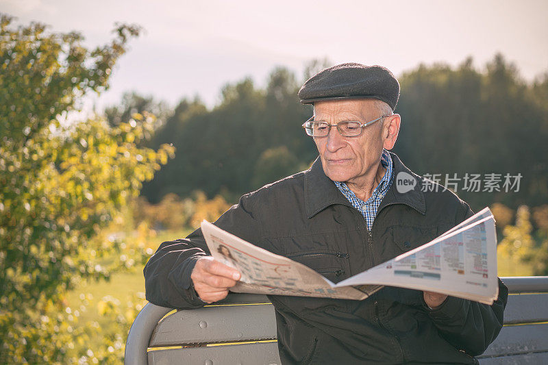 Elderly man wearing cap and glasses reading newspaper sitting on wooden bench in city public park